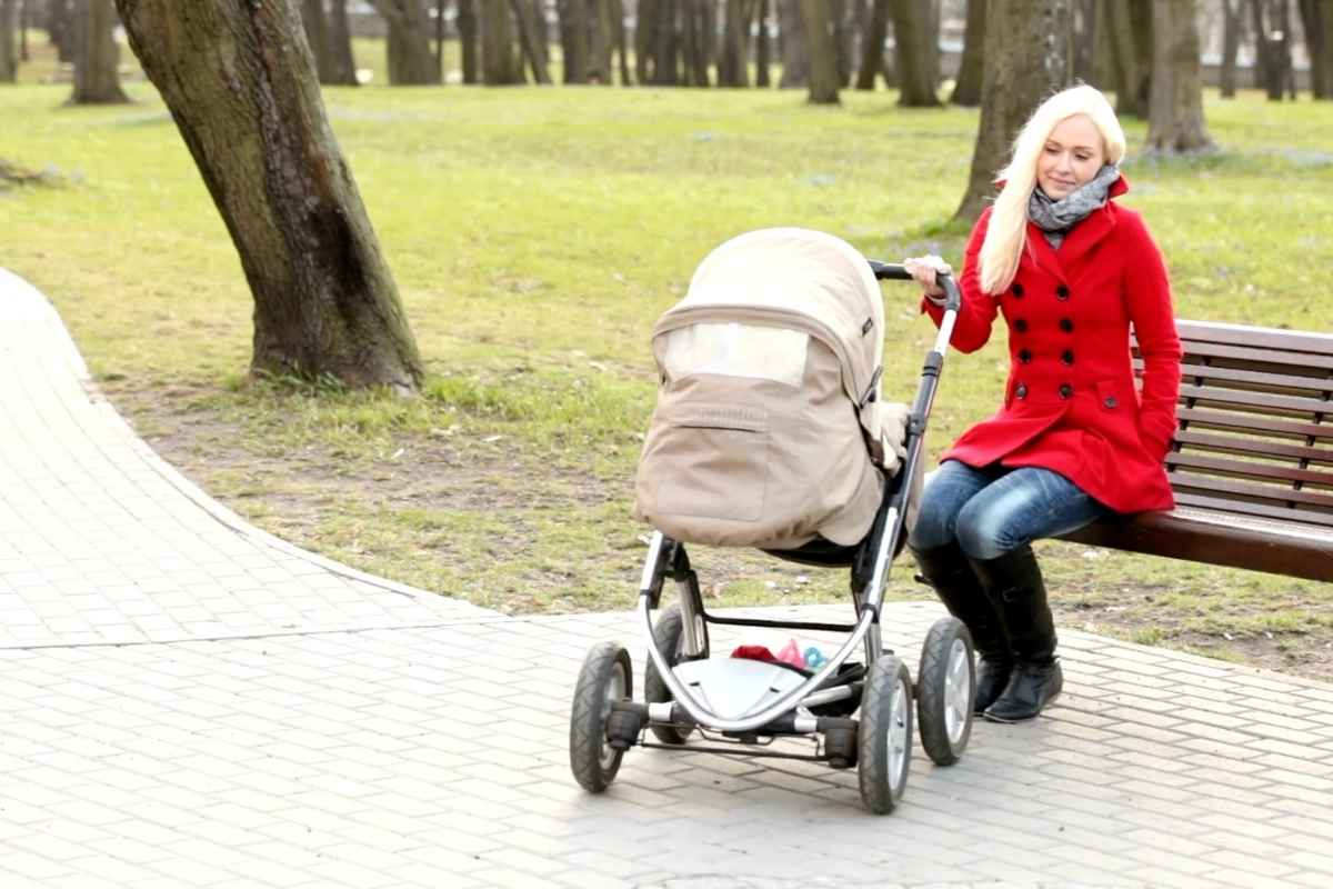 mama u crvenom kaputu sedi na klupici i gura bebu u kolicima